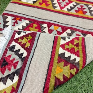 May include: A traditional handwoven rug with a geometric pattern in shades of red, white, black, and green. The rug has a fringed edge and is folded in half.