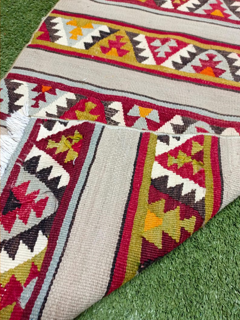 May include: A traditional handwoven rug with a geometric pattern in shades of red, white, black, and green. The rug has a fringed edge and is folded in half.