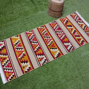 May include: A long, rectangular rug with a geometric pattern in shades of red, yellow, and white. The rug has a fringe on both ends.