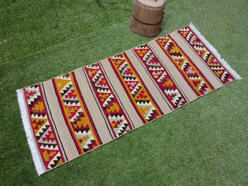 May include: A long, rectangular rug with a geometric pattern in shades of red, yellow, and white. The rug has a fringe on both ends.