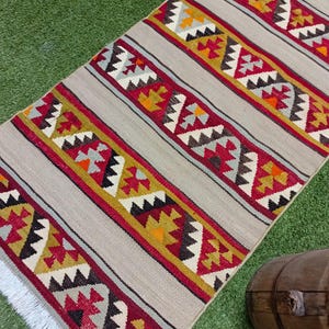 May include: A long, rectangular rug with a white background and colorful geometric patterns in shades of red, yellow, orange, and gray. The rug has a fringe on the bottom edge.
