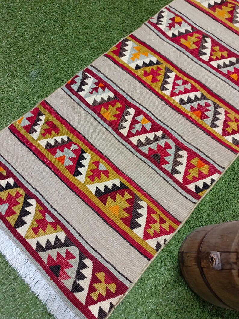 May include: A long, rectangular rug with a white background and colorful geometric patterns in shades of red, yellow, orange, and gray. The rug has a fringe on the bottom edge.