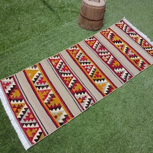 May include: A long, rectangular rug with a geometric pattern in shades of red, yellow, and black on a beige background. The rug has white fringe on the edges.