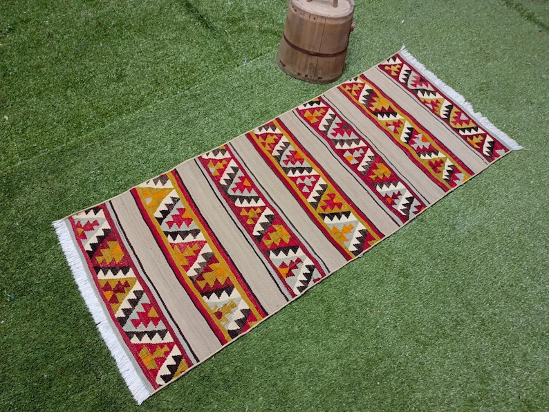 May include: A long, rectangular rug with a geometric pattern in shades of red, yellow, and black on a beige background. The rug has white fringe on the edges.
