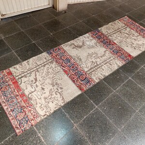 Puede incluir: Una alfombra rectangular de estilo vintage con un campo central beige y bordes decorativos. La alfombra presenta un patrón repetitivo con acentos rojos, azules y beige. La alfombra está colocada sobre un suelo de baldosas oscuras.