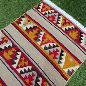 May include: A handwoven rug with a geometric pattern in shades of red, yellow, blue, and black on a beige background. The rug has a white fringe on the edges.