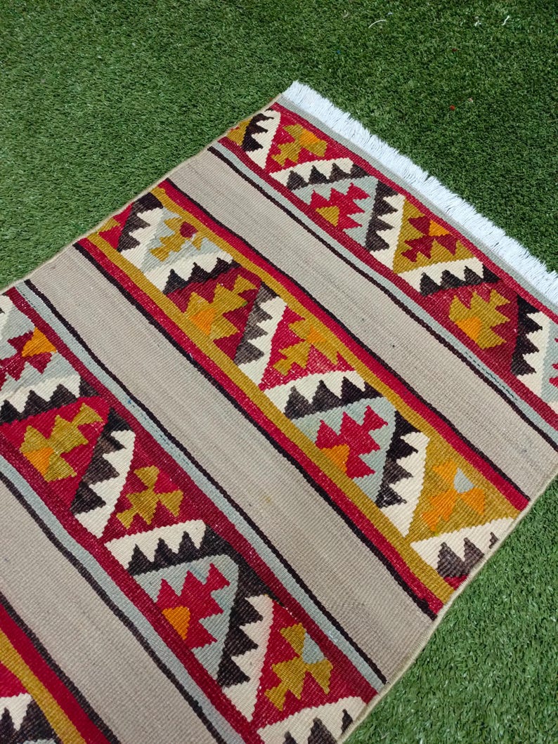 May include: A handwoven rug with a geometric pattern in shades of red, yellow, blue, and black on a beige background. The rug has a white fringe on the edges.