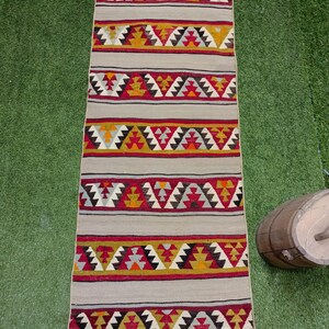 May include: A long, narrow rug with a beige background and colorful geometric patterns in shades of red, yellow, black, and white. The rug has a fringe on both ends.