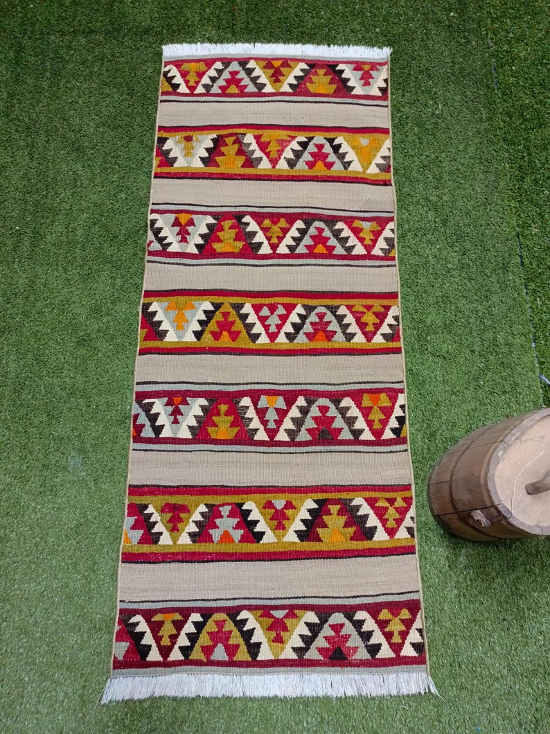 May include: A long, narrow rug with a beige background and colorful geometric patterns in shades of red, yellow, black, and white. The rug has a fringe on both ends.
