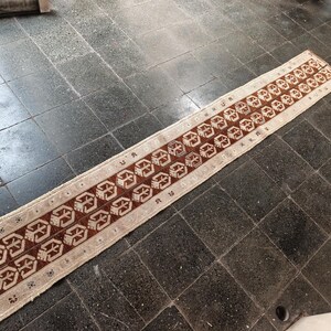 Puede incluir: Una alfombra de pasillo larga y rectangular con una base de color crema y un patrón geométrico repetitivo en marrón oscuro. El diseño consta de dos filas paralelas de motivos simétricos estilizados, terminada con flecos en ambos extremos.