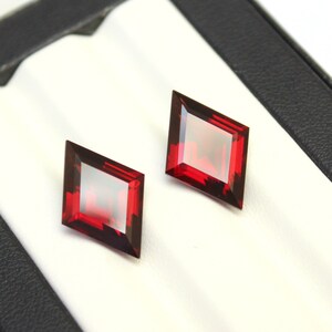 May include: Two red gemstone diamonds in a white display box. The gemstones are cut in a square shape with beveled edges.