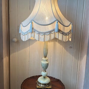 Peut inclure: Une lampe de sol vintage avec un abat-jour cr&egrave;me, des glands bleus et une base d&eacute;corative. La lampe a un corps vert clair et dor&eacute; et repose sur une petite table ronde en bois. La hauteur de la lampe est d'environ 150 cm.