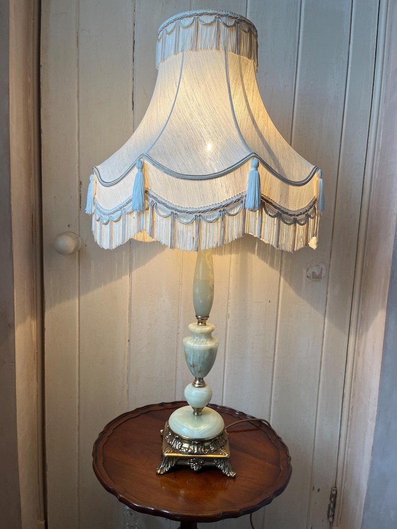 Peut inclure: Une lampe de sol vintage avec un abat-jour cr&egrave;me, des glands bleus et une base d&eacute;corative. La lampe a un corps vert clair et dor&eacute; et repose sur une petite table ronde en bois. La hauteur de la lampe est d'environ 150 cm.
