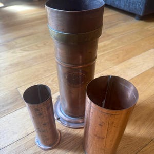 May include: Three copper cylinders of different sizes. The largest has a wide top and a brass band. The middle cylinder has a circular base and an embossed emblem. The smallest is tapered. All are on a wooden surface.