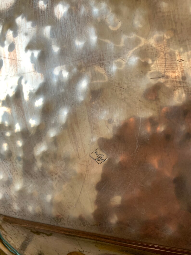 Pu&ograve; includere: Primo piano di un vassoio d'argento con un marchio a forma di diamante al centro. Il marchio contiene un uccello stilizzato con le ali spiegate.