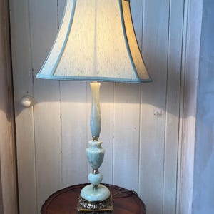 Peut inclure: Une lampe de table vintage avec un abat-jour bleu clair et cr&egrave;me. La lampe a une base et une tige orn&eacute;es vert clair et repose sur une table ronde marron fonc&eacute;. La base de la lampe est carr&eacute;e avec des accents dor&eacute;s d&eacute;coratifs.
