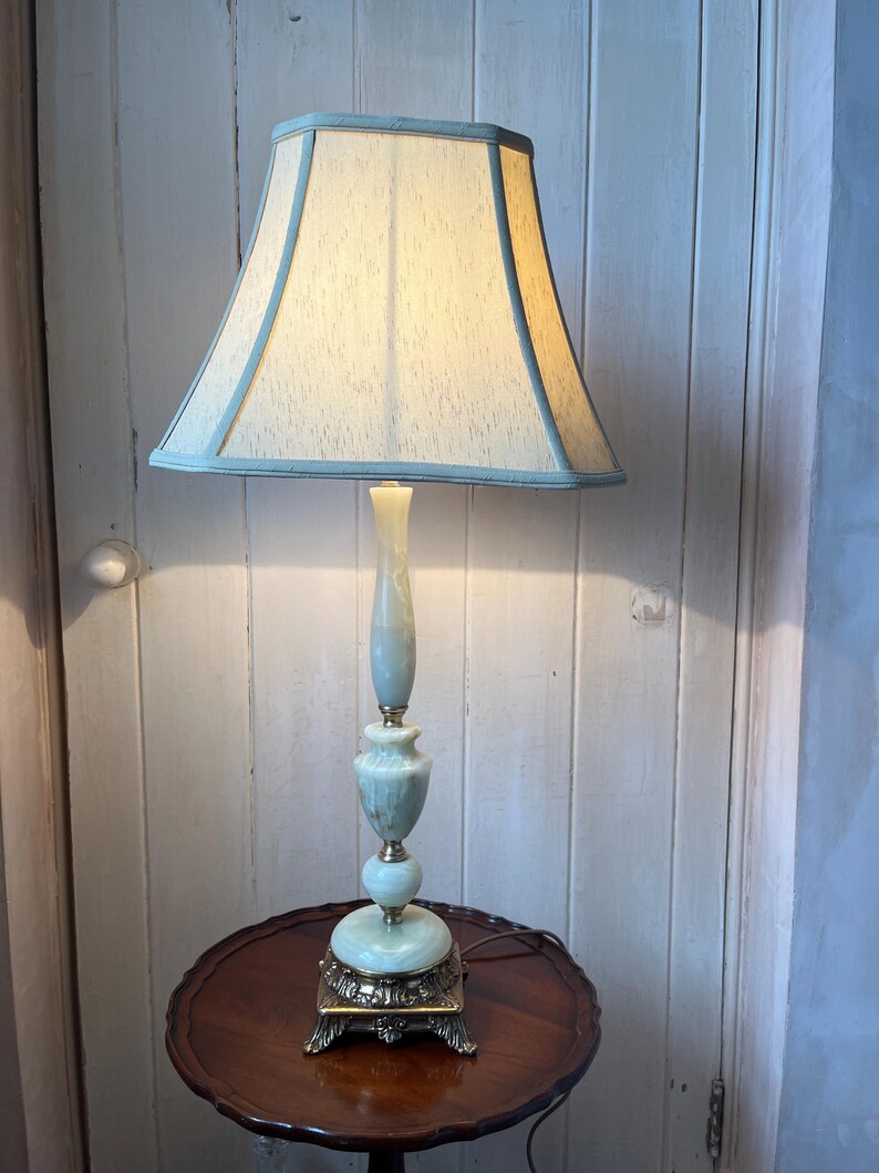 Peut inclure: Une lampe de table vintage avec un abat-jour bleu clair et cr&egrave;me. La lampe a une base et une tige orn&eacute;es vert clair et repose sur une table ronde marron fonc&eacute;. La base de la lampe est carr&eacute;e avec des accents dor&eacute;s d&eacute;coratifs.
