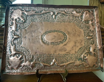 May include: A copper serving tray with a hammered finish and an embossed floral design. The tray has two handles and a raised oval in the center.