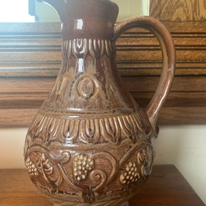 May include: A brown ceramic pitcher with a textured surface and a grapevine design. The pitcher has a handle and a spout. The word "Vino" is embossed on the front of the pitcher.
