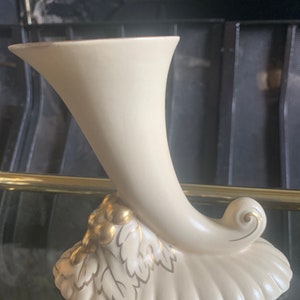 Op de afbeelding: Een crèmekleurige keramische vaas in de vorm van een hoorn met gouden accenten. De vaas heeft een bladontwerp en een tros gouden druiven aan de basis.
