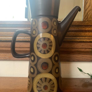 May include: A tall brown ceramic coffee pot with a geometric pattern in shades of brown, beige, and red. The pot has a lid and a handle.