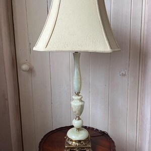 Peut inclure: Une lampe de table vintage avec un abat-jour cr&egrave;me et une base vert clair et dor&eacute;e. La lampe est pos&eacute;e sur une table ronde marron fonc&eacute;. La base de la lampe pr&eacute;sente un design orn&eacute; et d&eacute;coratif.