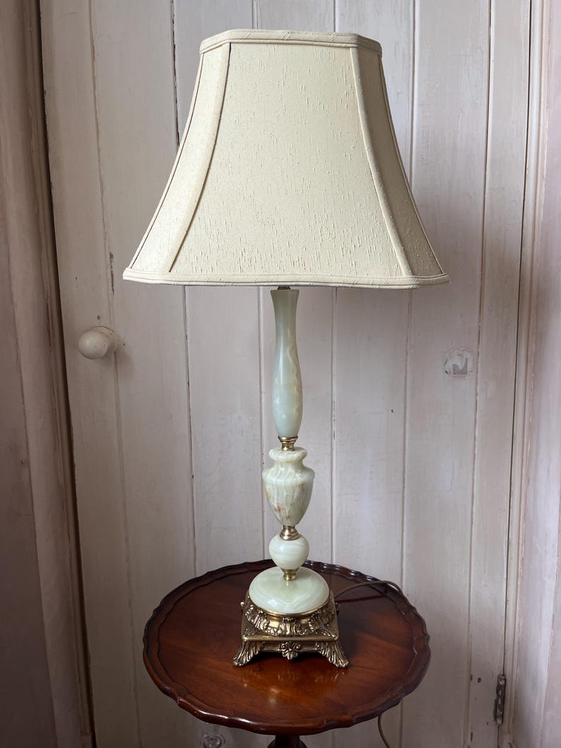 Peut inclure: Une lampe de table vintage avec un abat-jour cr&egrave;me et une base vert clair et dor&eacute;e. La lampe est pos&eacute;e sur une table ronde marron fonc&eacute;. La base de la lampe pr&eacute;sente un design orn&eacute; et d&eacute;coratif.