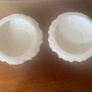 May include: Two white ceramic bowls with a fluted rim. The bowls are empty and sitting on a wooden surface.