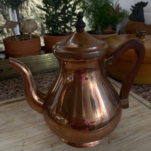 Puede incluir: Una tetera de cobre con asa de madera y un pomo de madera en la tapa. La tetera es brillante y refleja la luz.