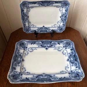 May include: Two rectangular, white porcelain serving platters with ornate blue floral and scrollwork borders. One platter is displayed on a black stand, showcasing the detailed design and elegant aesthetic of the vintage tableware.