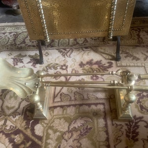 Puede incluir: Un juego de herramientas para chimenea de latón con una pala, pinzas y un atizador. Las herramientas están montadas en un soporte con un diseño decorativo.