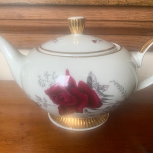Könnte beinhalten: Weiße Teekanne mit goldener Verzierung und einem roten Rosenmuster. Die Teekanne hat einen runden Korpus und einen Ausguss an der Seite.