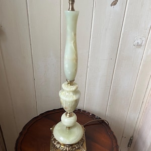 Peut inclure: Une lampe de table vintage avec un corps en onyx vert clair et des accents en laiton. La lampe pr&eacute;sente une base carr&eacute;e avec des d&eacute;tails orn&eacute;s, une partie centrale renfl&eacute;e et une colonne &eacute;lanc&eacute;e. La lampe mesure environ 61 cm de haut.