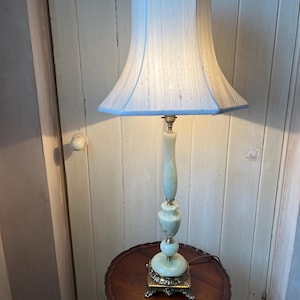 Peut inclure: Une lampe de table vintage avec un abat-jour bleu clair et une base vert p&acirc;le. La lampe a un pied en laiton et repose sur une petite table ronde en bois. L'abat-jour est de couleur bleu clair.