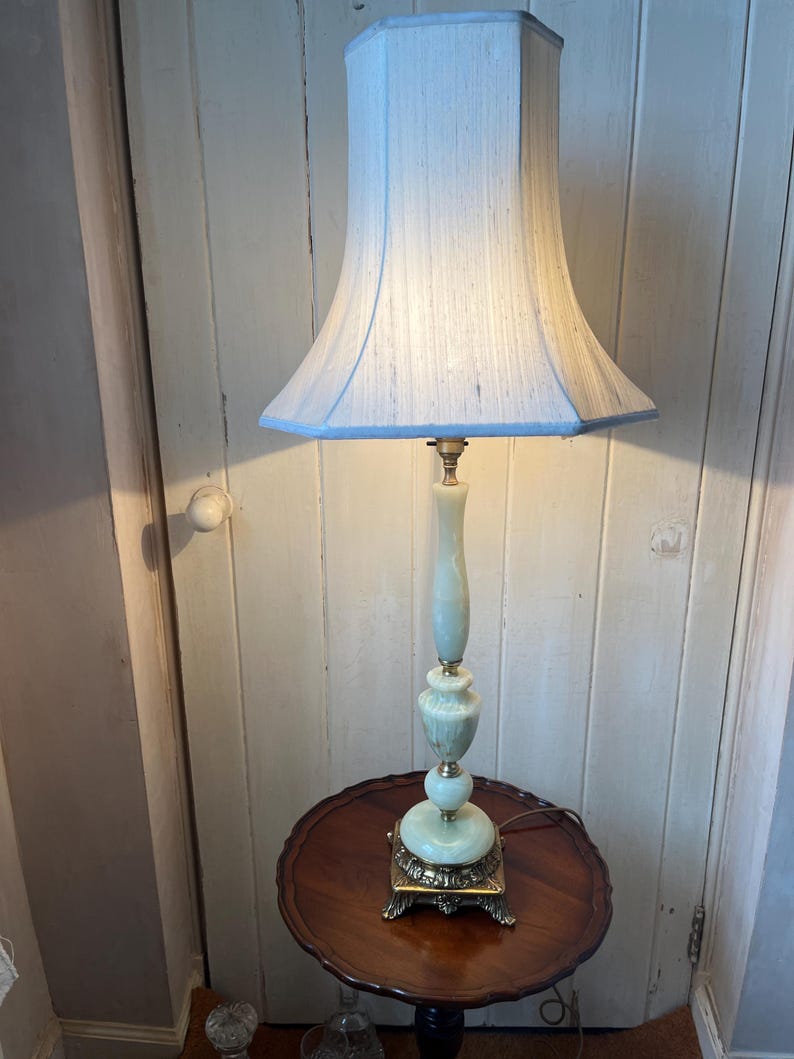 Peut inclure: Une lampe de table vintage avec un abat-jour bleu clair et une base vert p&acirc;le. La lampe a un pied en laiton et repose sur une petite table ronde en bois. L'abat-jour est de couleur bleu clair.