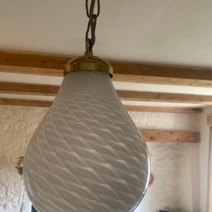 Könnte beinhalten: Eine weiße Glaspendelleuchte mit einem wirbelnden Muster hängt an einer Messingkette. Die Leuchte ist tropfenförmig und hat ein goldfarbenes Metalloberteil.