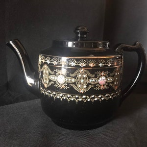 May include: Black teapot with gold and white decorative trim. The teapot has a round body and a spout on the left side. The handle is on the right side.