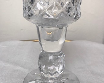 May include: Clear glass candle holder with a diamond pattern on the top and a round base. The candle holder has a short stem and is designed to hold a small candle. The glass is transparent.