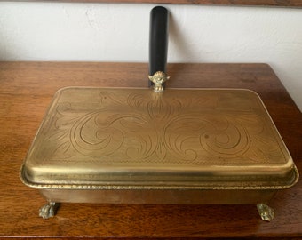 May include: A brass box with a decorative engraved design on the lid. The box has a black handle and four small feet.