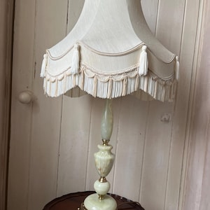 Peut inclure: Une lampe de table vintage avec un abat-jour cr&egrave;me orn&eacute; de glands et de franges. La lampe pr&eacute;sente une base et une tige en marbre vert et blanc, et une base d&eacute;corative en m&eacute;tal dor&eacute;. La lampe mesure environ 76 cm de haut.