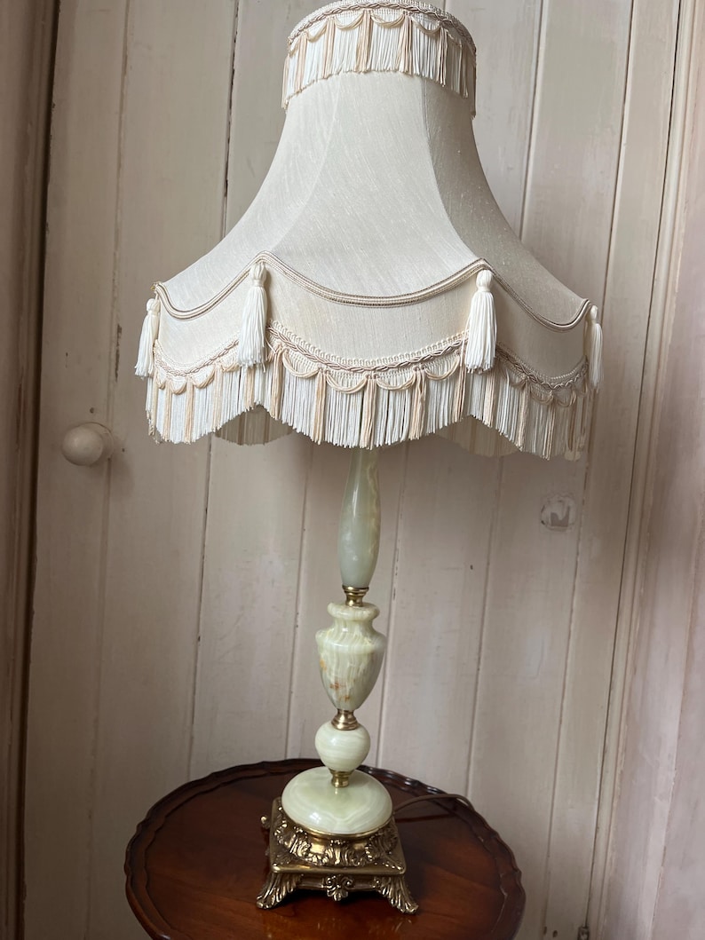 Peut inclure: Une lampe de table vintage avec un abat-jour cr&egrave;me orn&eacute; de glands et de franges. La lampe pr&eacute;sente une base et une tige en marbre vert et blanc, et une base d&eacute;corative en m&eacute;tal dor&eacute;. La lampe mesure environ 76 cm de haut.