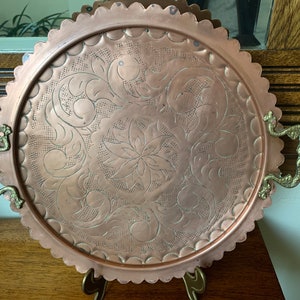 Può includere: Un vassoio rotondo in rame per servire con un bordo a festoni e un design floreale decorato. Il vassoio ha due manici in ottone.