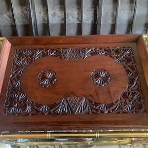 Può includere: Un vassoio da portata in legno marrone scuro con intricati disegni intagliati. Il vassoio ha un bordo rialzato e due disegni circolari al centro.