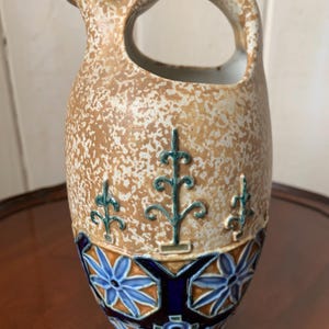 Può includere: Brocca in ceramica con corpo screziato beige e crema, un grande manico e un beccuccio. La base presenta motivi floreali blu e arancioni. Accenti verdi stilizzati a forma di albero sono posti sul corpo.