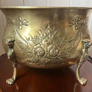 Può includere: Una fioriera in ottone con dettagli floreali e fiocchi. La fioriera ha tre piedini decorativi a zampa di leone. L'esterno presenta rose in rilievo, foglie e altri elementi floreali. La fioriera è di colore oro caldo.