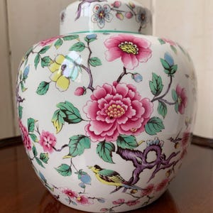 May include: A white ceramic ginger jar with a lid, adorned with a floral pattern featuring pink peonies, yellow and pink flowers, green leaves, and a yellow bird. The jar has a round shape.