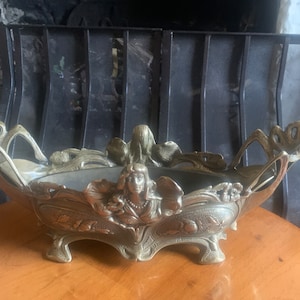 May include: An antique brass-toned centerpiece bowl with Art Nouveau styling. The bowl features a central female figure and floral motifs. The bowl is supported by three feet and has handles on each side. The bowl is displayed on a wooden table.