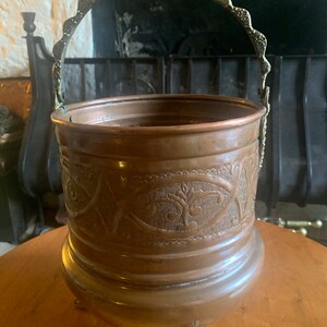 May include: An antique copper bucket with an ornate brass handle and three decorative feet. The bucket has an embossed design around the middle.