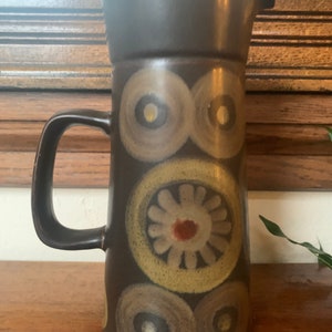 May include: A brown ceramic pitcher with a geometric pattern of circles and flowers in shades of brown, yellow, and white.