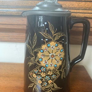 May include: Black ceramic pitcher with a pewter lid and a floral design in gold, blue, and white. The pitcher has a handle and a spout.